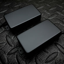Two black guitar humbucker pickups with gold-toned screw terminals, displayed on a textured dark gray surface.