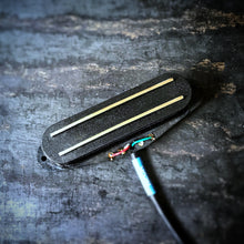 A close-up of a black guitar single coil sized humbucker pickup with two rails and a black cable, resting on a dark textured surface.