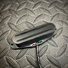 A close-up of a black guitar pickup with a twin rails, resting on a textured gray diamond plate metal surface.