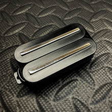 A close-up of a black double rail metal guitar pickup with two silver metal pole pieces, resting on a textured black diamond plate background.